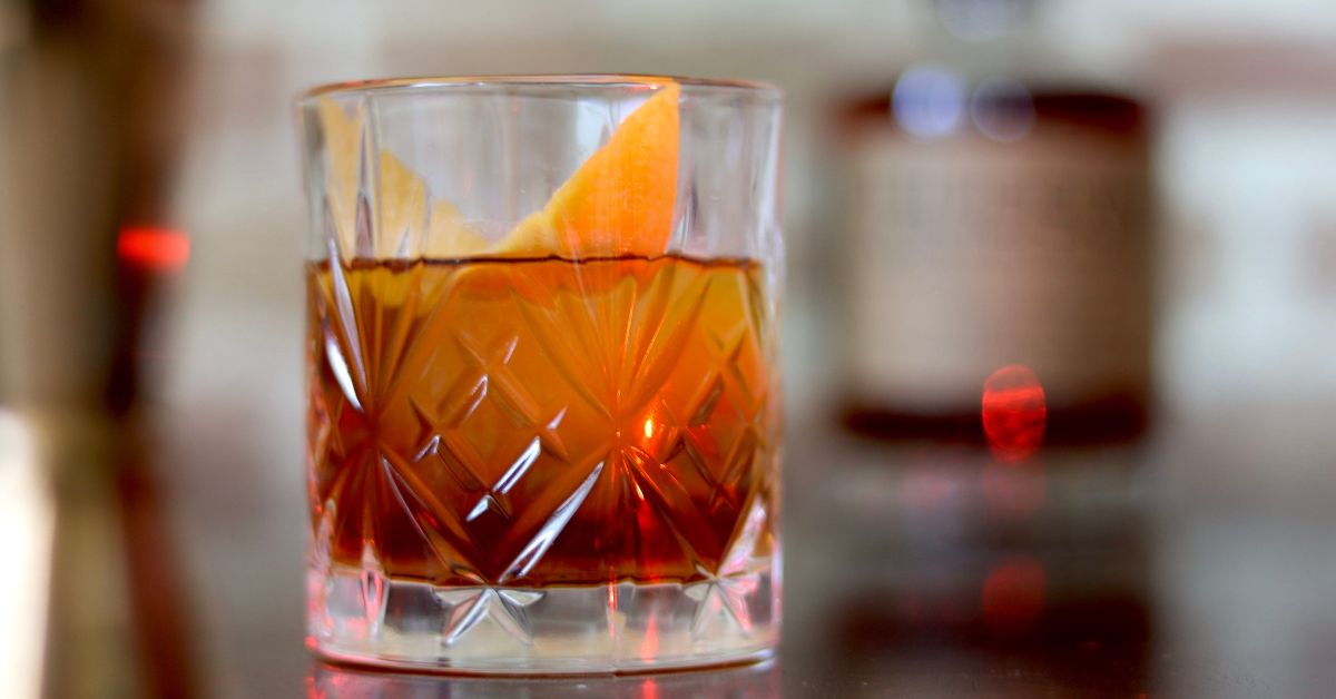 An Old Fashioned in a crystal glass, garnished with an orange peel, sits in focus on a bar top, with a whiskey bottle out of focus in the background.